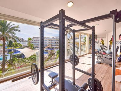 Helles Fitnessstudio mit Aussicht auf Pool und Palmen durch große Fenster.