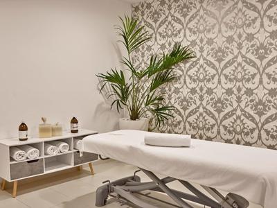 Ein ruhiger Massageraum mit weißem Massagebett, Handtüchern und einer grünen Pflanze vor gemusterter Tapete.