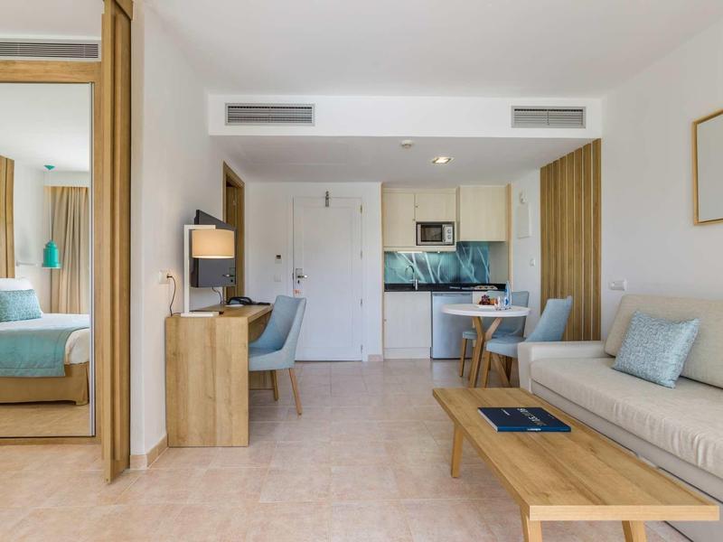 Habitación de hotel moderna y luminosa con área de estar, escritorio, cocina pequeña y dormitorio separado.
