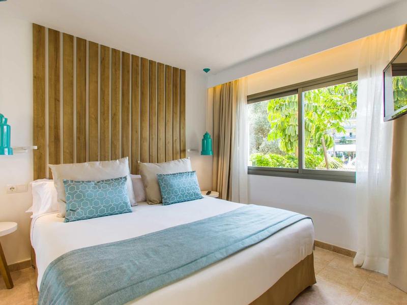 Dormitorio luminoso con cama doble, acentos azules y vista al verde a través de gran ventana.