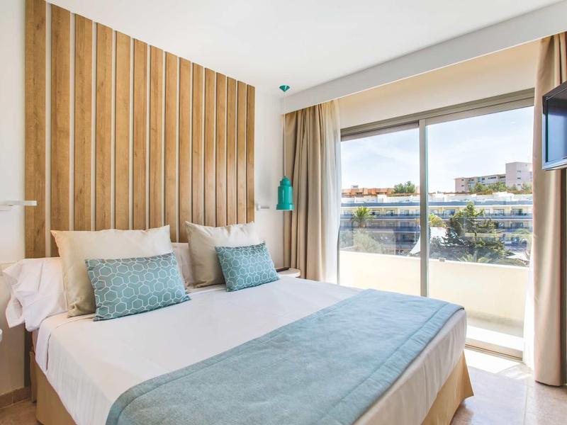 Habitación de hotel luminosa con cama doble, ropa de cama azul y blanca y balcón con vista a la ciudad.