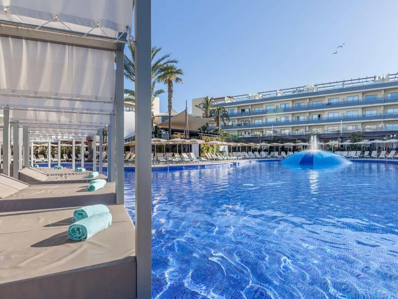 Piscina moderna de hotel con agua azul, tumbonas y palmeras bajo cielo despejado.
