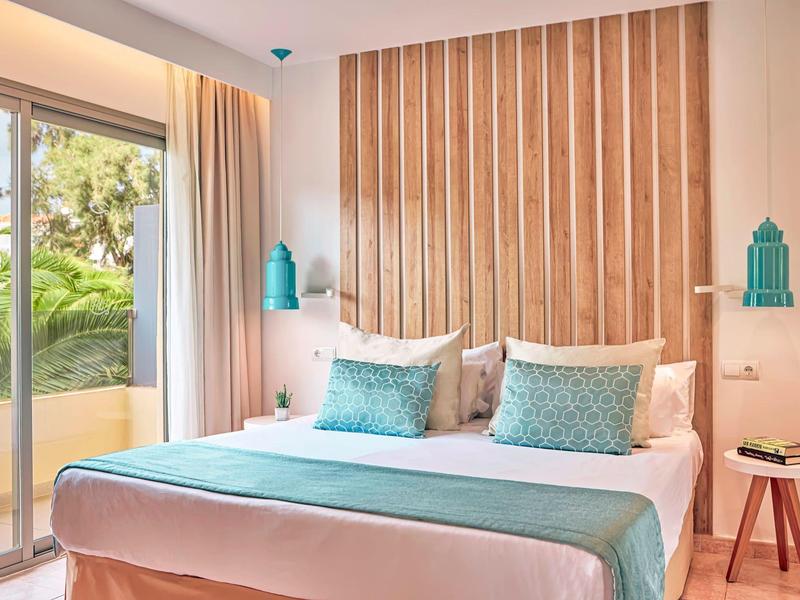 Helles Hotelzimmer mit Doppelbett, türkisfarbenen Akzenten und Balkon mit Pflanzenblick.