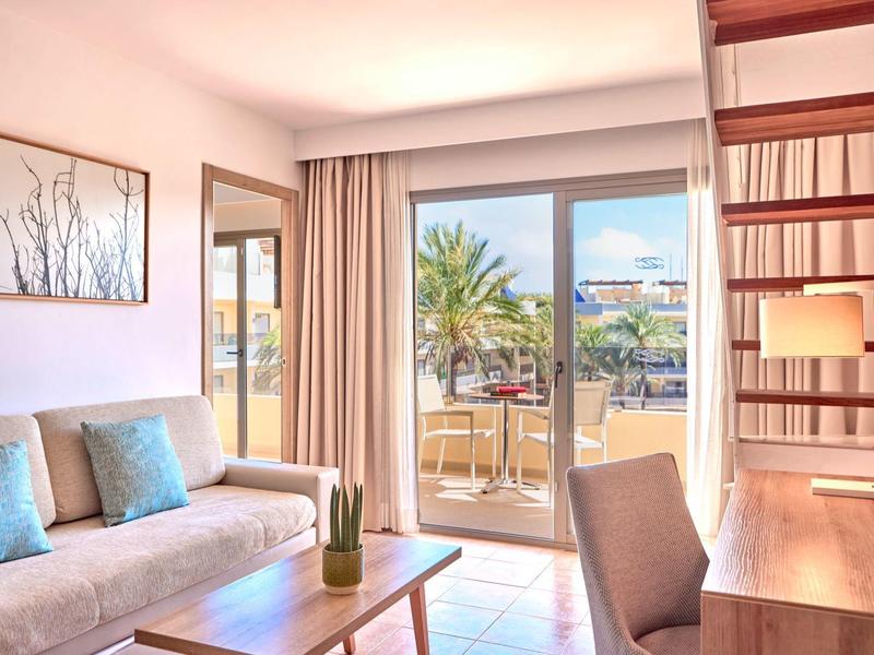 Helles Hotelzimmer mit Sofa, Holztisch, Schreibtisch und Blick auf Terrasse mit Palmen.