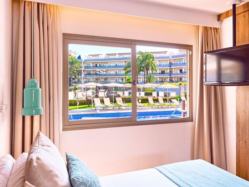 Blick aus einem Hotelzimmer auf einen Poolbereich mit Liegestühlen und Palmen.