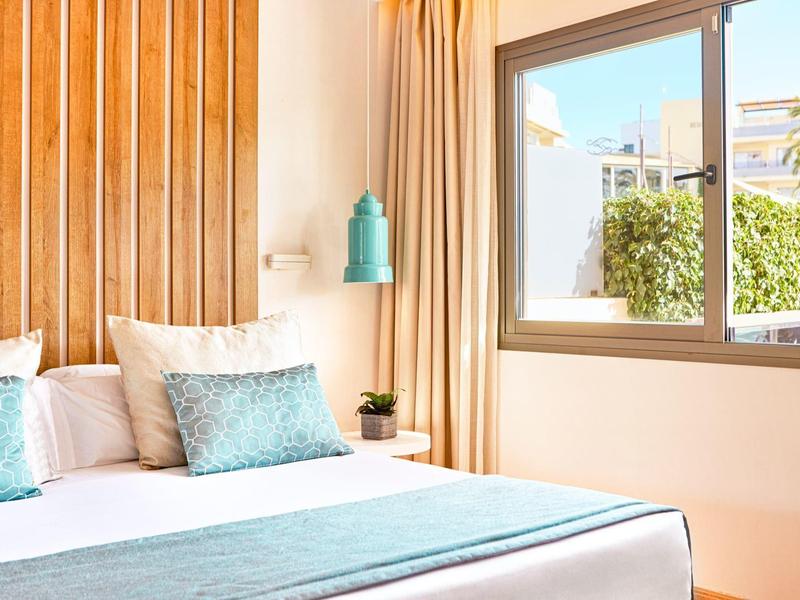 Hell beleuchtetes Schlafzimmer mit Holzdekoration, großem Fenster und blauen Akzenten im Kissen und Bettdecke.