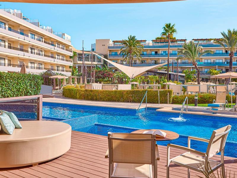 Moderner Hotelpool mit Sonnenliegen, Palmen und mehrstöckigen Gebäuden im Hintergrund.