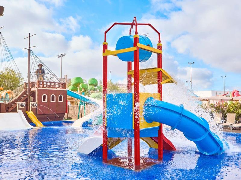 Wasserpark mit blauen Rutschen, einem Piratenschiff und spritzendem Wasser unter blauem Himmel.