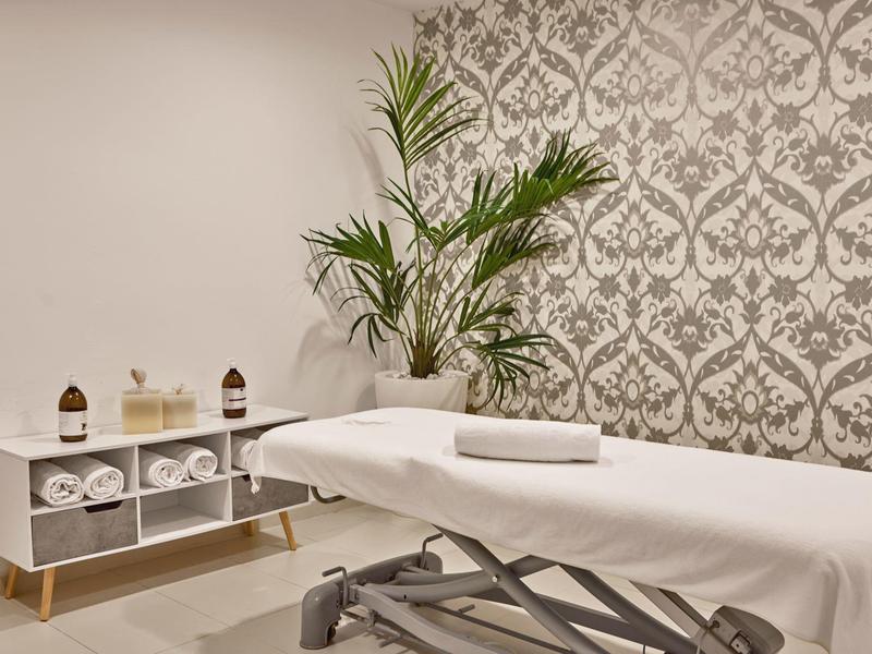 Ein ruhiger Massageraum mit weißem Massagebett, Handtüchern und einer grünen Pflanze vor gemusterter Tapete.