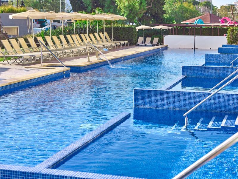 Großer Außenpool mit blauen Fliesen, Wasserdüsen und Liegestühlen im Hintergrund.