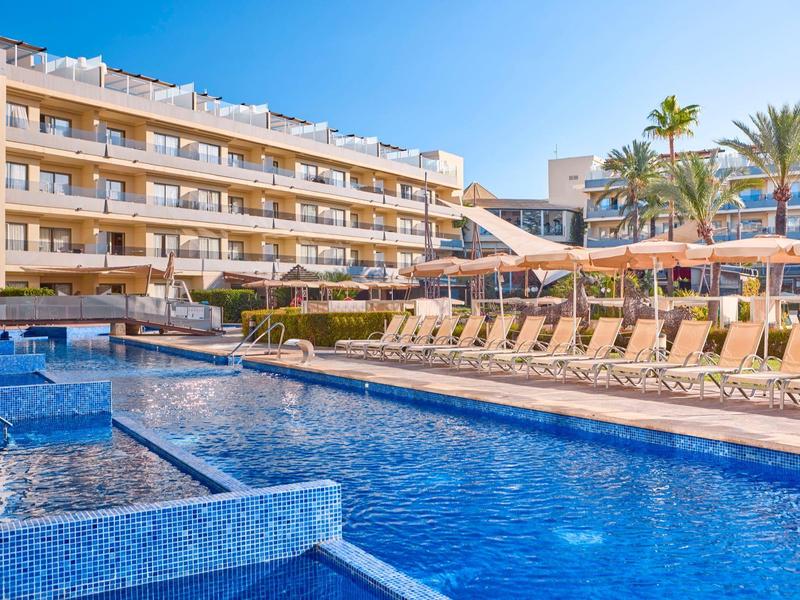 Großer Pool vor einem fünfstöckigen Hotel mit Liegestühlen und Sonnenschirmen.