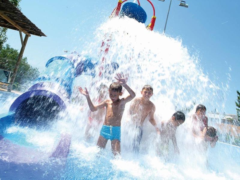 Niños jugando y riendo mientras agua de una regadera grande cae en parque acuático.