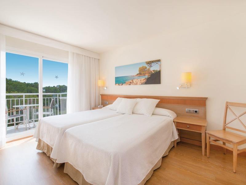 Jasny pokój hotelowy z łóżkiem podwójnym, balkonem i widokiem na zielone wzgórza.