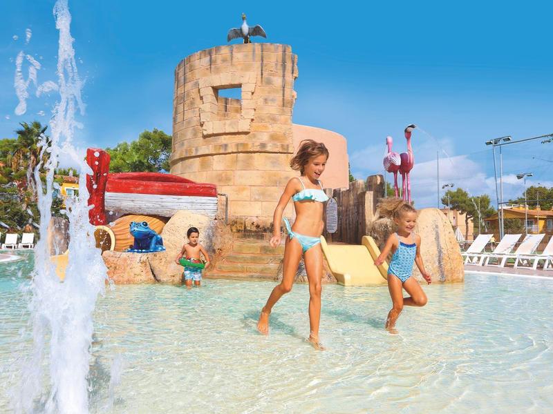 Dzieci bawiące się w płytkim basenie z atrakcjami wodnymi i zamkiem w tle.
