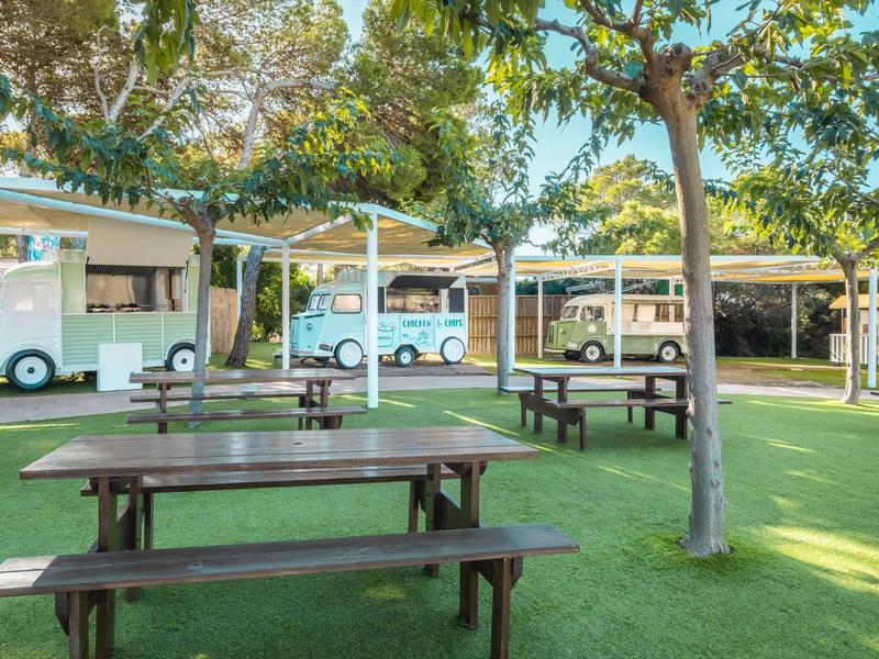 Zielony teren z ławkami i stołami pod drzewami obok kamperów.