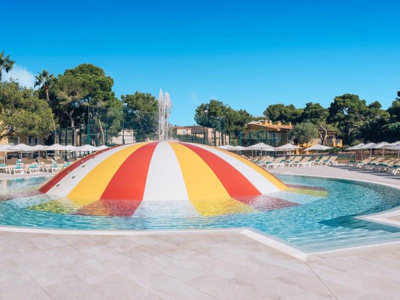 Kolorowy basen z fontanną i leżakami pod niebieskim niebem.