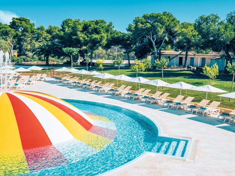 Duży basen hotelowy z fontanną i leżakami w słonecznym ogrodzie.