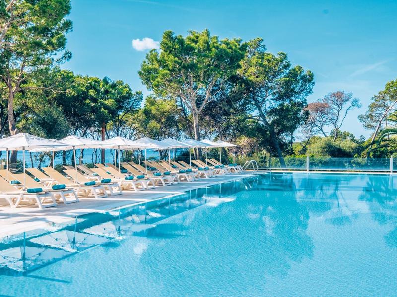 Duży odkryty basen z leżakami i parasolami w słonecznym hotelu otoczonym drzewami.