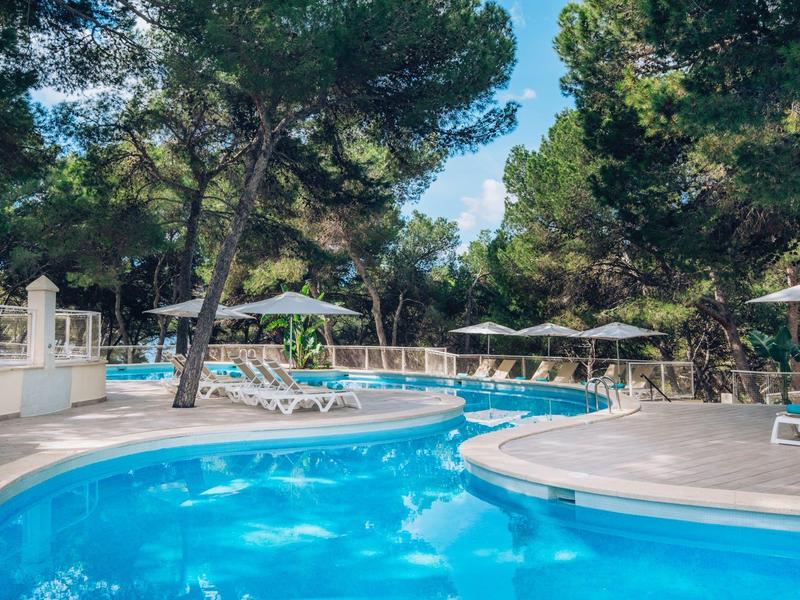 Okrągły basen hotelowy otoczony leżakami i wysokimi sosnami w słoneczny dzień.