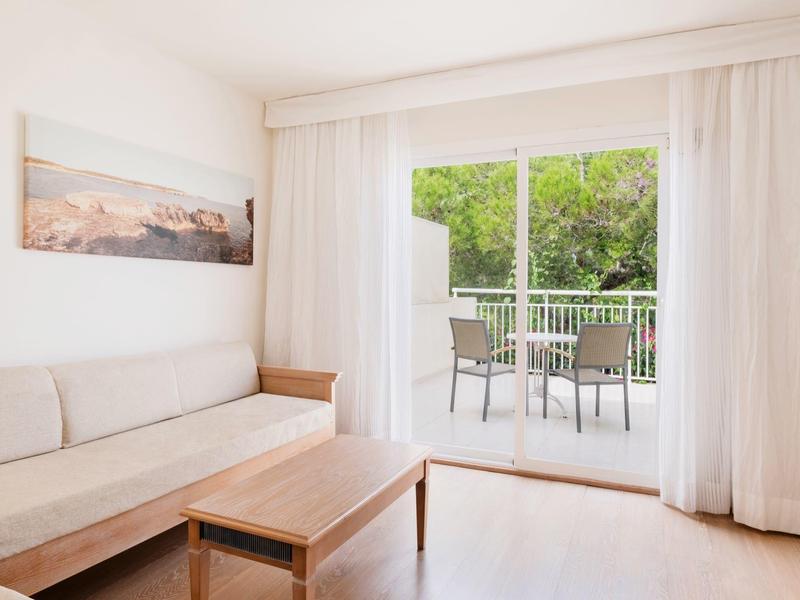 Jasny pokój hotelowy z sofą, drewnianym stołem, balkonem z krzesłami i widokiem na zieleń.