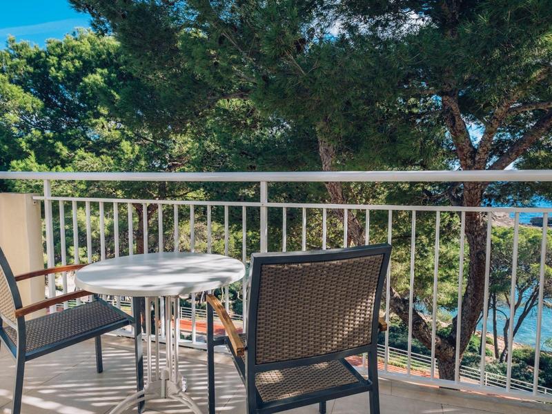 Balkon z okrągłym stołem i dwoma krzesłami, widok na drzewa i morze.