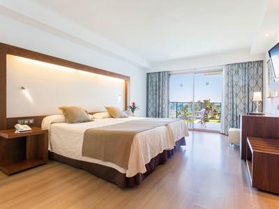 Bright hotel room with a large bed, wooden furniture, and balcony overlooking greenery.