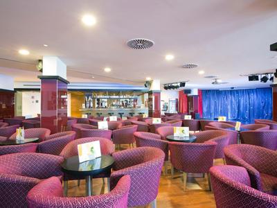 Spacious hotel lounge with purple armchairs, small round tables, and a bar in the background.