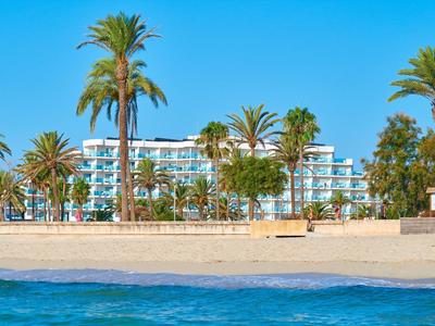 Hotel with palm trees on a sandy beach next to clear blue sea under a bright sky.