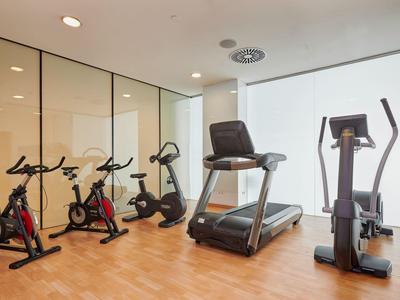 Modern gym room with stationary bikes, treadmill, and elliptical on wooden floor.