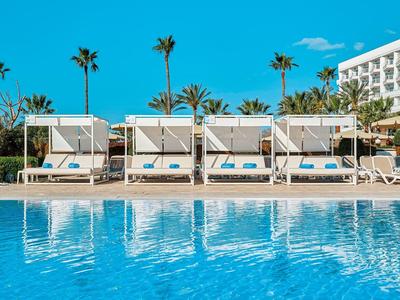 Luxury pool loungers with sunshades in front of a hotel, surrounded by palm trees and clear blue sky.