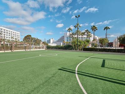 Artificial turf soccer field with palm trees surrounded by hotel buildings under a blue sky