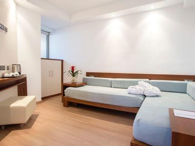 Modern hotel room with corner sofa, desk, and light wooden furniture.