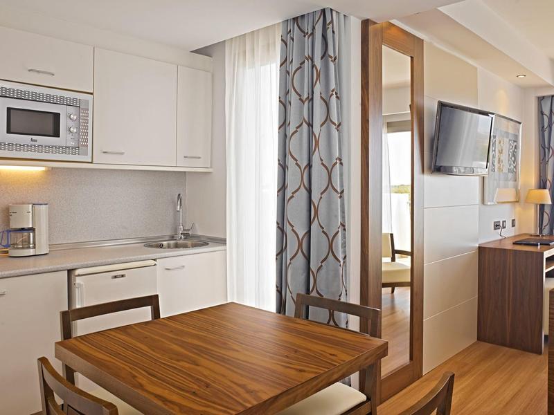 Bright hotel room with kitchenette, wooden dining table, chairs, and a wall-mounted TV.