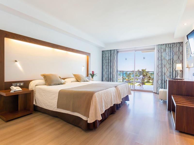 Bright hotel room with a large bed, wooden furniture, and balcony overlooking greenery.