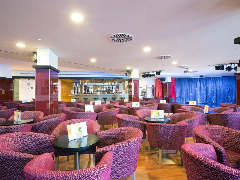 Spacious hotel lounge with purple armchairs, small round tables, and a bar in the background.