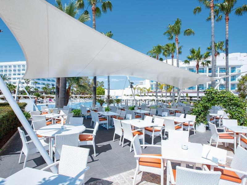 Outdoor hotel dining area with white tables and chairs under sunshades surrounded by palm trees.