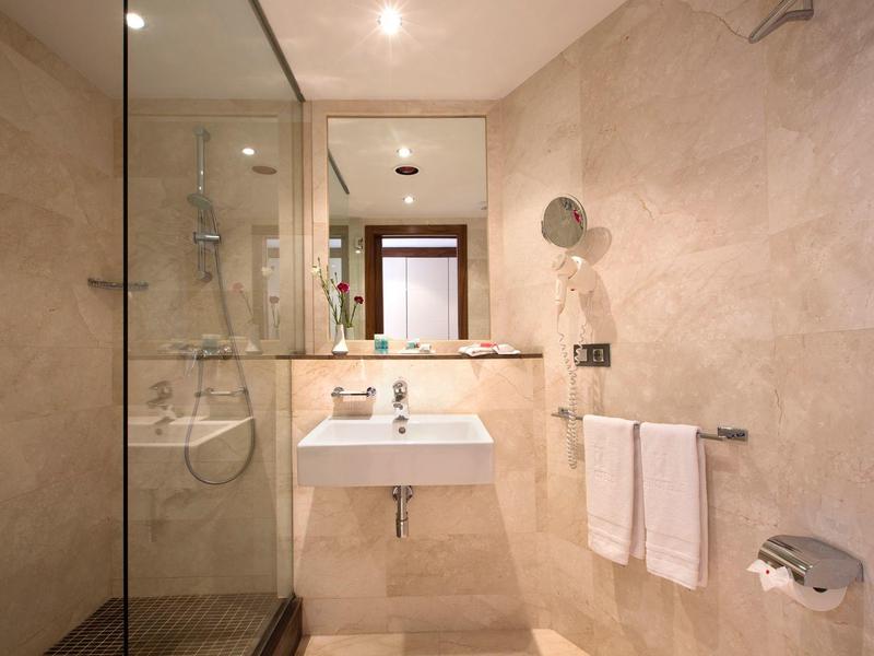 Modern hotel bathroom with glass shower, white sink, large mirror, and beige tiled walls.