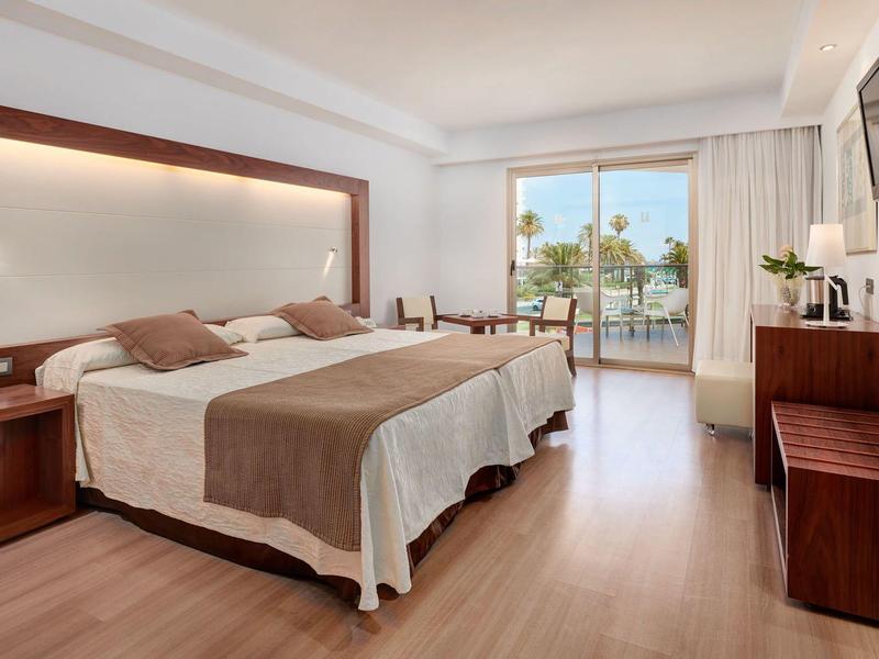 Bright hotel room with double bed, wooden furniture, and balcony overlooking palm trees.