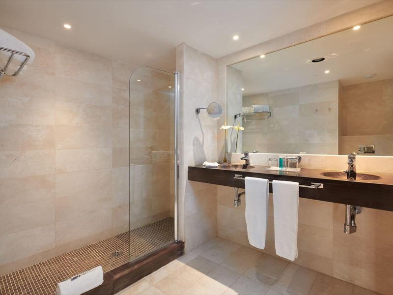 Modern bathroom with glass shower, large mirror, double sink, and beige tiled walls.