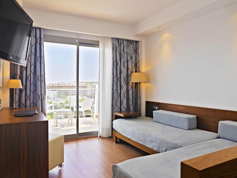 Bright hotel room with two single beds, TV, wooden furniture, and balcony with city view.