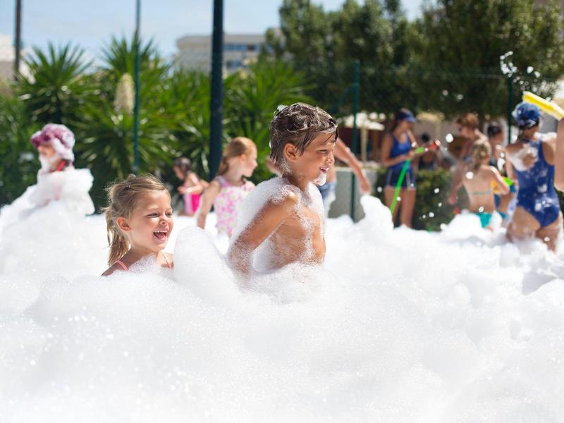Children playing and enjoying a large foam party outdoors on a sunny day near palm trees.
