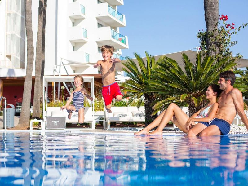 Two children jump into a pool while two adults sit by the poolside on a sunny day.