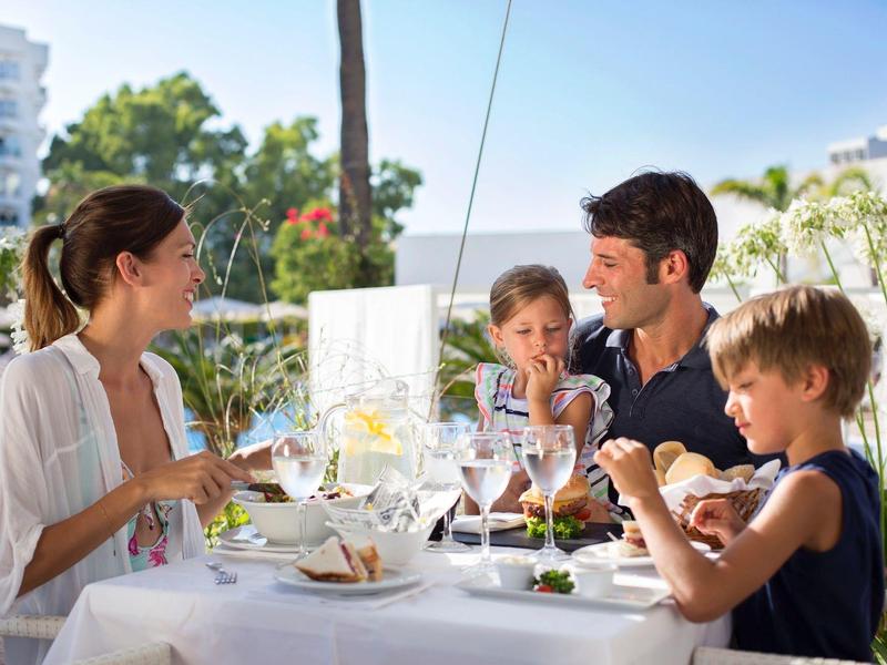 Family enjoying meal on terrace with pool in background.
