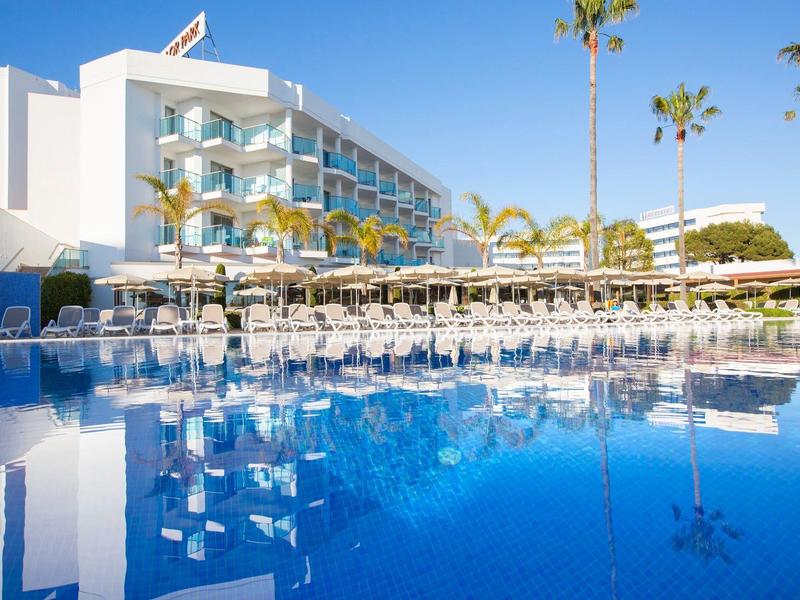 Modern hotel complex with pool, sun umbrellas, and palm trees on a sunny day.