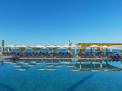 Grande area piscina dell'hotel con lettini e ombrelloni bianchi sotto un cielo azzurro limpido.