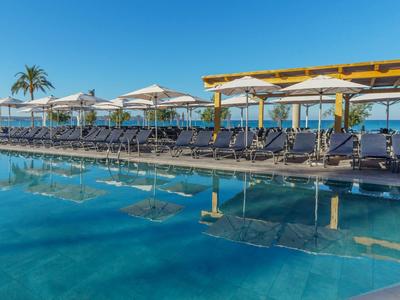 Grande piscina con lettini e ombrelloni vicino al mare sotto un cielo limpido.