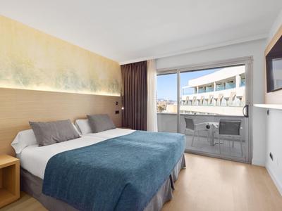 Camera d'hotel moderna con letto matrimoniale, balcone e vista sugli edifici circostanti.