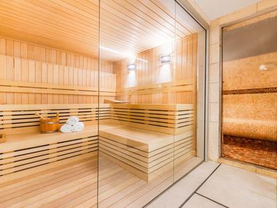 Sauna luminosa con panchine in legno e parete in vetro, a fianco bagno di vapore con luce calda.