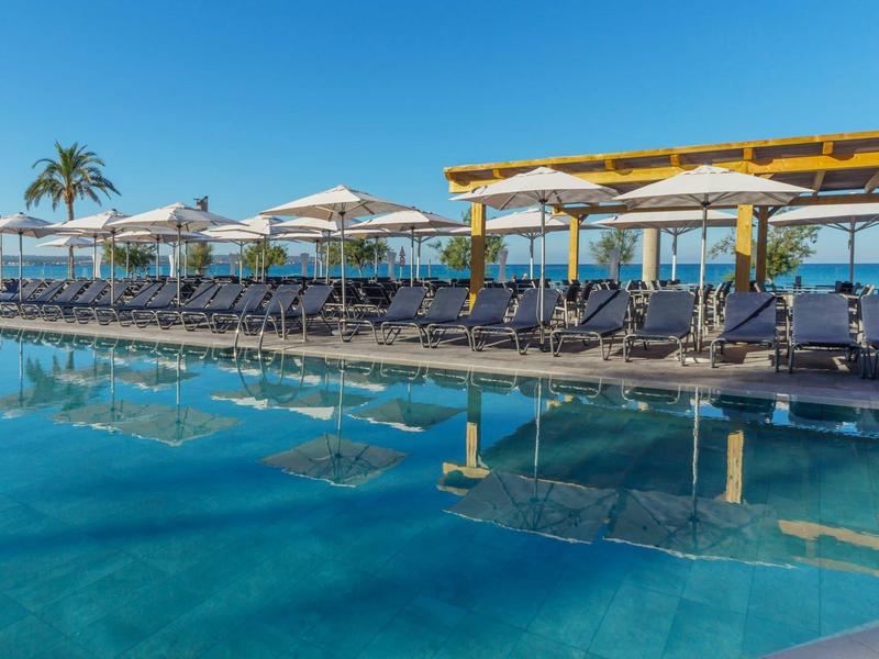 Grande piscina con lettini e ombrelloni vicino al mare sotto un cielo limpido.