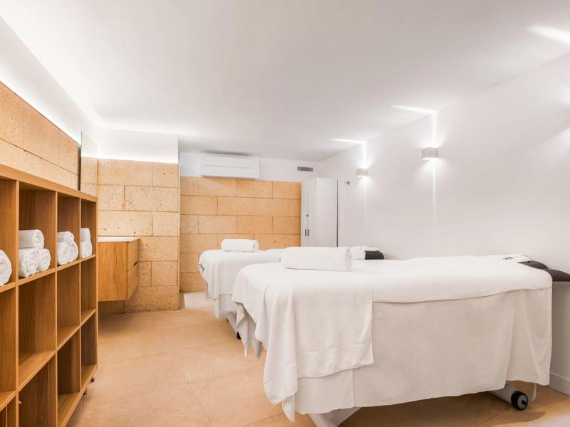 Studio massaggi moderno e luminoso con lettini bianchi e scaffale in legno con asciugamani.